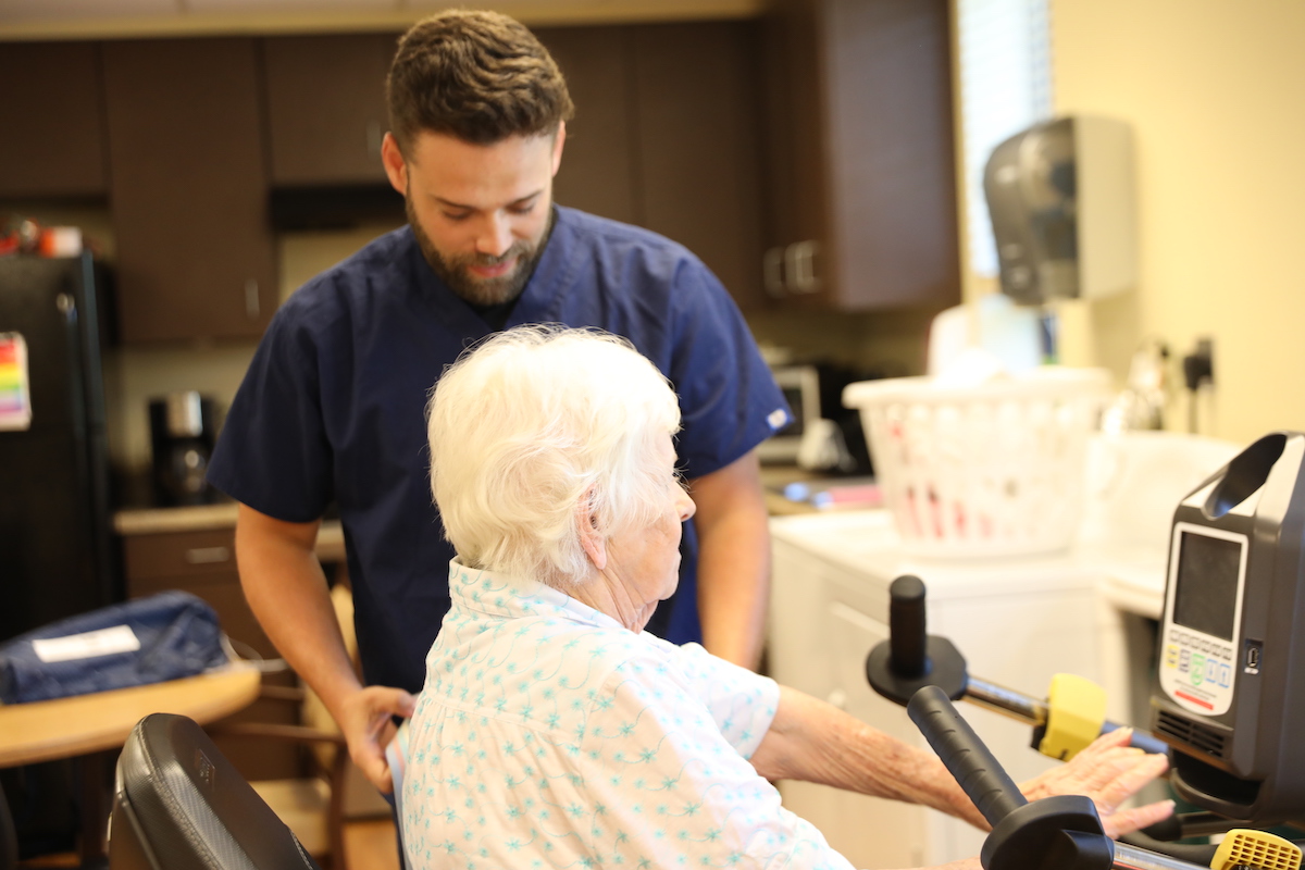 Male Caregiver Assists Senior Woman Perform Exercises on Stationary Bike_Uplands Village