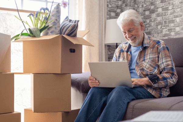 Happy senior man involved in moving house sitting among cardboard boxes using laptop, concept of relocating, retirement, new life, buying, renting, apartment, house