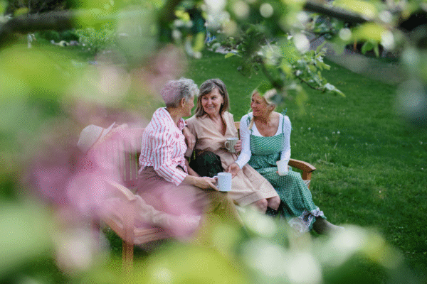 Happy senior women friends sitting on bench and drinking tea outdoors in garden, laughing.