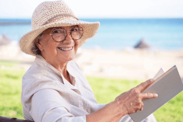 Smiling senior woman relaxes on a sunny beach while reading a book, wearing a straw sun hat and glasses. Calm, leisurely moment conveying retirement, relaxation, vacation and joyful outdoor living.