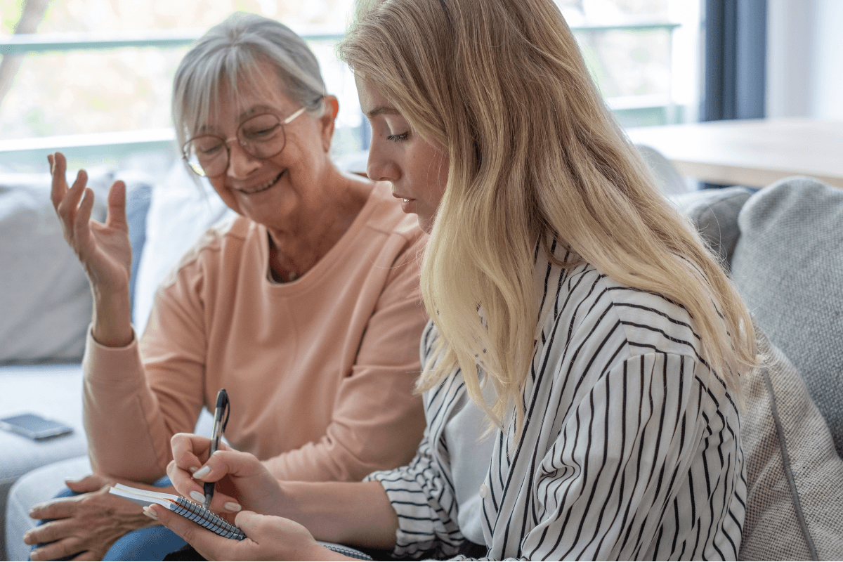 Mother and daughter sit on a sofa sharing a warm conversation while the younger woman takes notes in a notebook, capturing family stories, planning, and caregiving moments in a cozy living room.