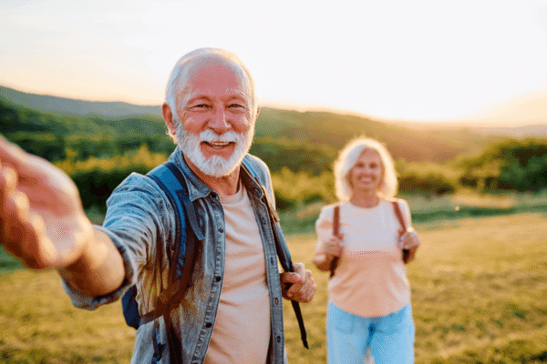 Senior man smiling at camera holding hand out while hiking with his wife, enjoying nature and active retirement.